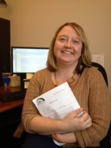 Anne Kelsey sits holding a packet of paper, smiling, with her computer screen behind her shoulder