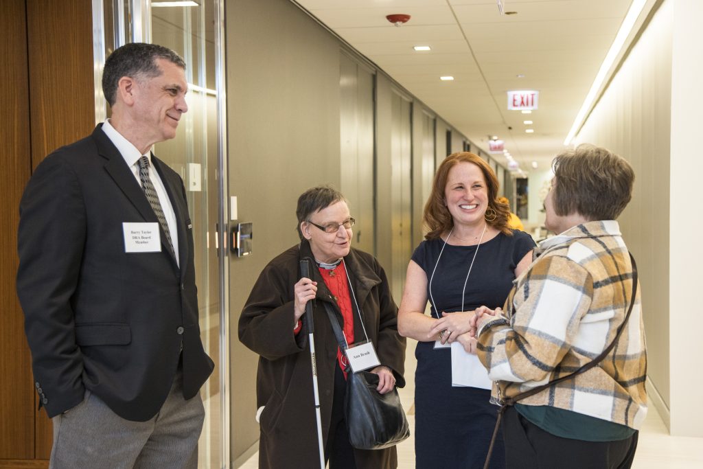 Four people including DRA Board member Barry Taylor and DRA's Rachel Weisberg smiling and speaking with each other in an office hallway. One of the guests is holding her white cane.