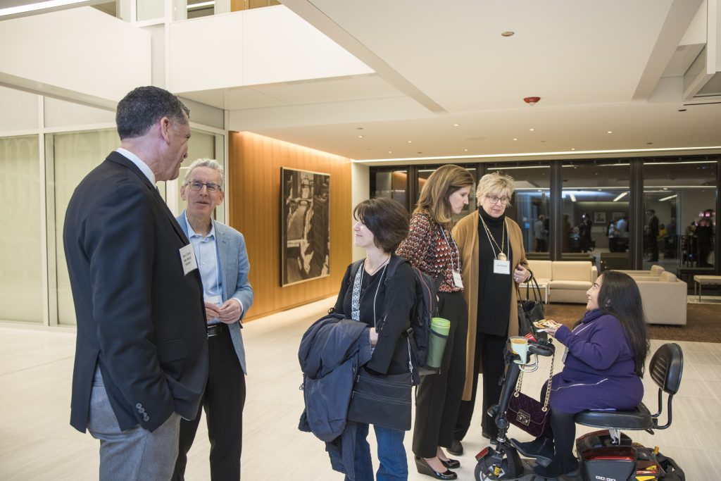 DRA Board member Barry Taylor speaks with two guests while three other guests speak separately. One of the guests is using her mobility scooter. Large windows, empty couches, and a large picture on a wall can be seen behind them.