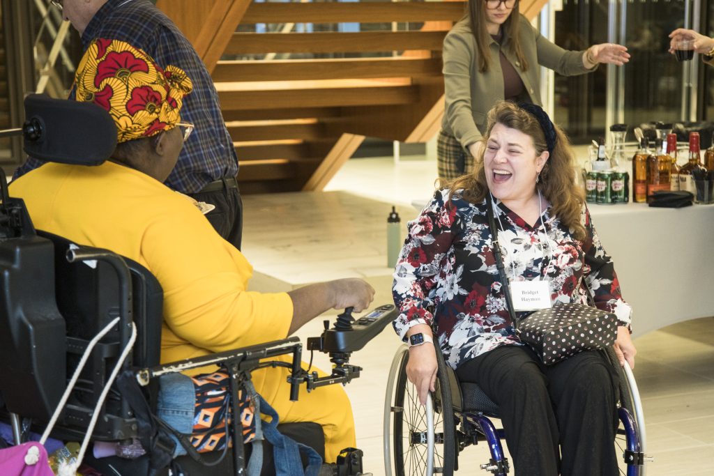 A guest using her wheelchair smiling as she greets another guest also using her wheelchair. Another guest and a large wooden staircase can be seen behind them.