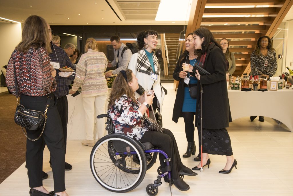 Four guests smiling and talking. One of the guests is using her wheelchair. Two other guests are speaking nearby. Other people and a wooden staircase can be seen behind them.