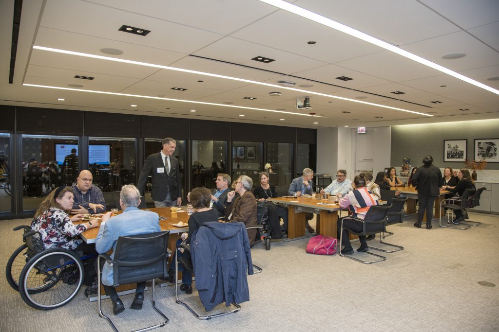19 guests gathered around three separate tables talking with each other, including DRA Board member Barry Taylor and DRA's Emily Roznowski. Most guests are sitting, but some are standing and some are using wheelchairs. Large windows are in the background.