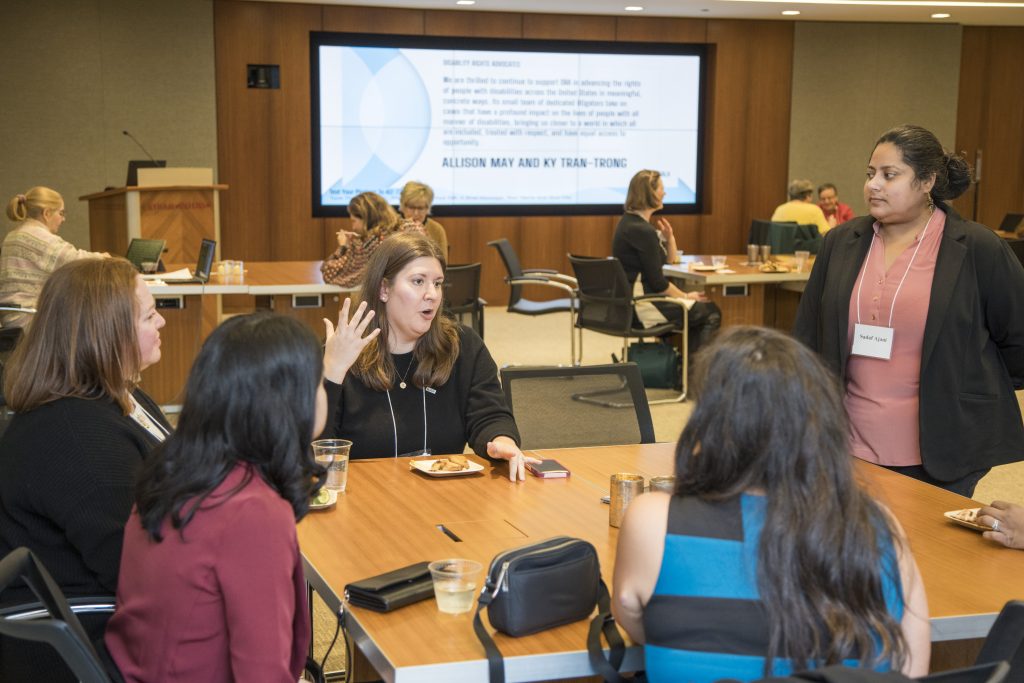 DRA's Emily Roznowski using her hand to gesture as she speaks with four guests gathered around a table. One guest is standing and the others are sitting. Other guests are sitting at tables in the background. A quote from Allison May and Ky Tran-Trong is displayed on a screen.