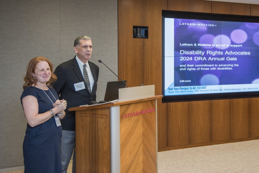 DRA's Rachel Weisberg smiling and standing next to DRA Board member Barry Taylor at a podium as Barry speaks into a microphone. A screen displays a message of support from sponsors Latham & Watkins.