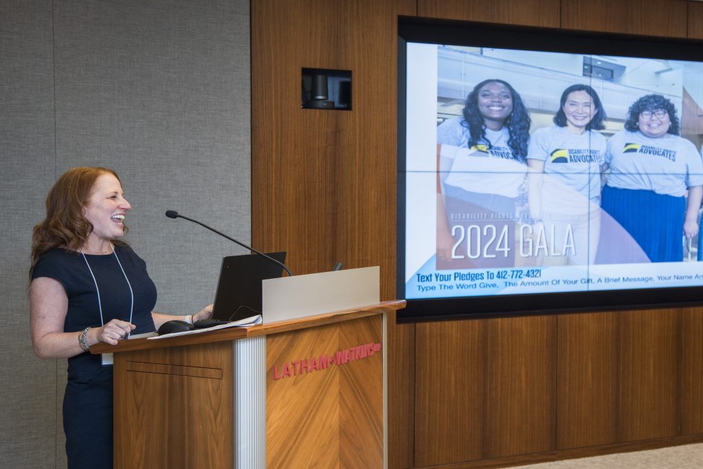 DRA's Rachel Weisberg at a podium smiling and speaking into a microphone. A screen displays DRA's Jameelah Najieb, DRA's Kinny Kim, and former DRA intern Sullyari Bautista.