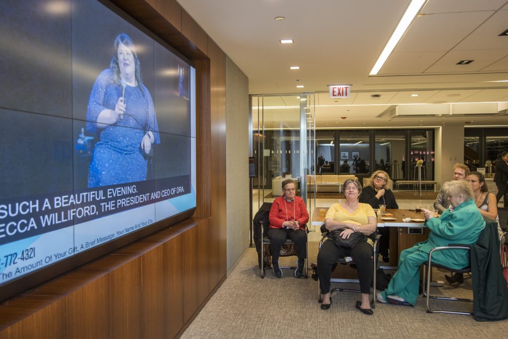 Six guests seated at a table watching the on-screen program that is currently displaying DRA's Rebecca Williford. Large windows are in the background.