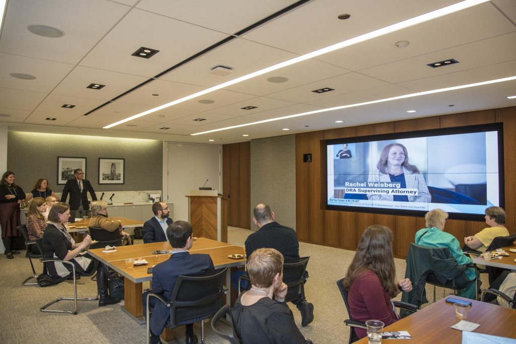 Many guests seated at tables watching the on-screen program that is currently displaying DRA's Rachel Weisberg.