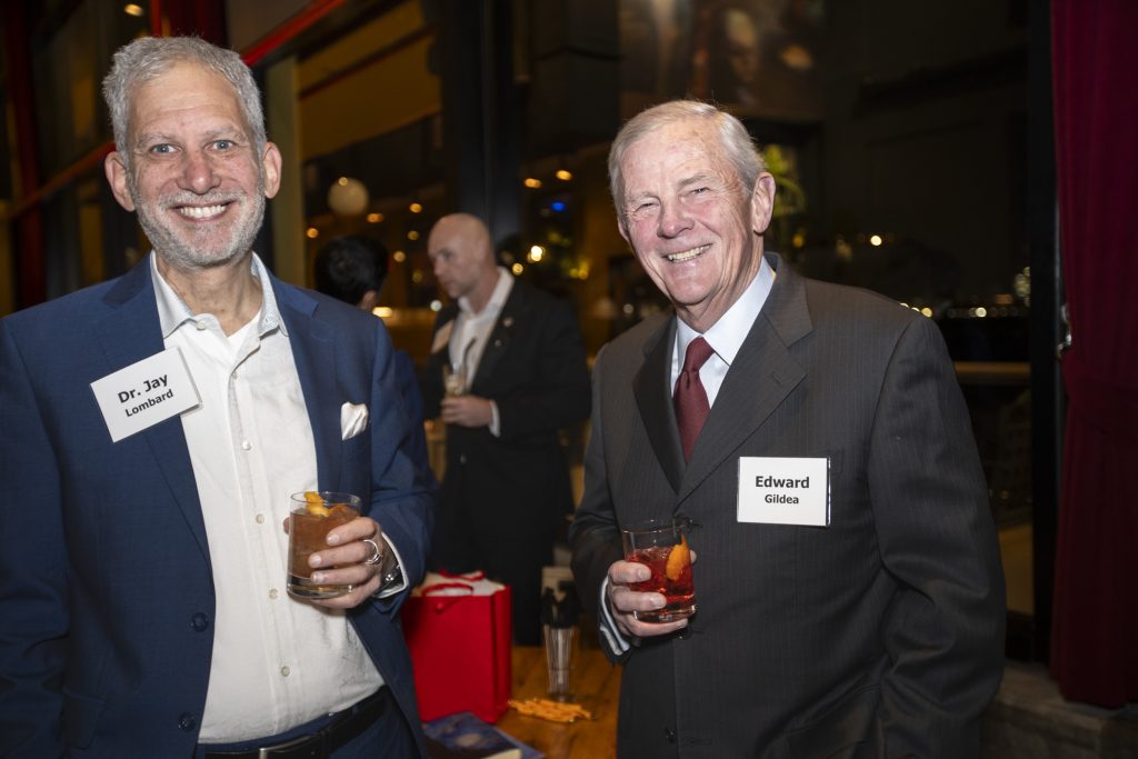 DRA Advisory Board member Edward Gildea posing with a guest, both smiling. Another guest can be seen behind them and city lights can be seen through the window.