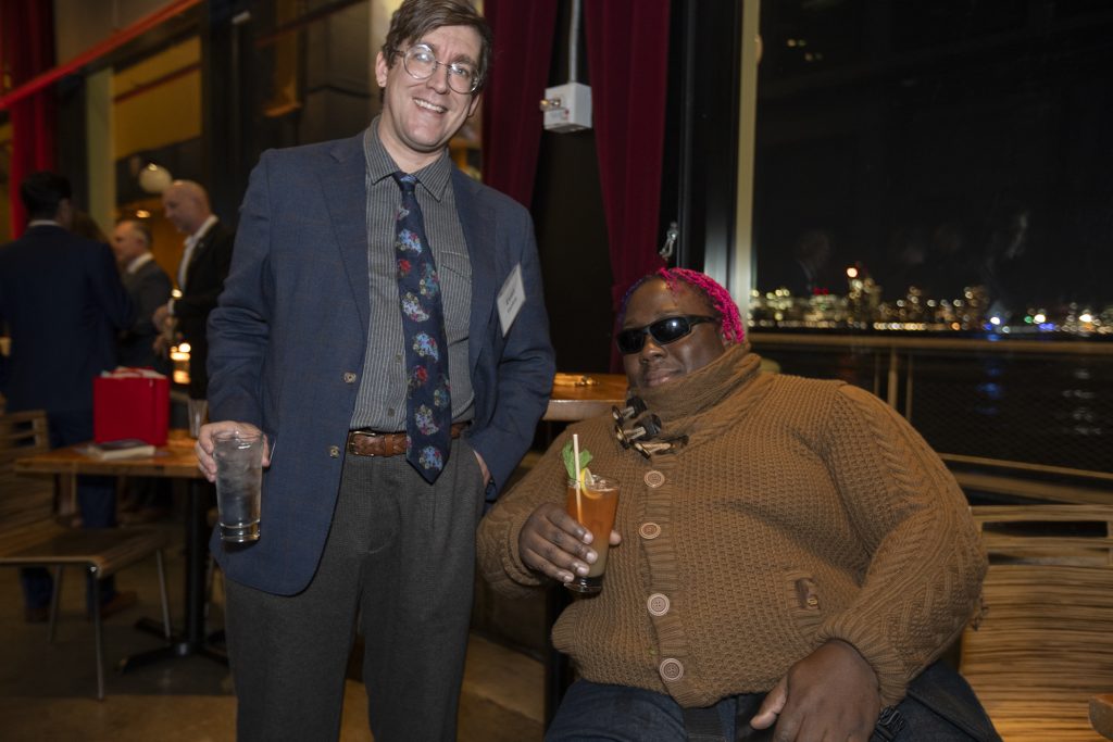 Two guests posing and smiling. One guest is using a wheelchair. Other guests can be seen behind them and city lights can be seen through the window.