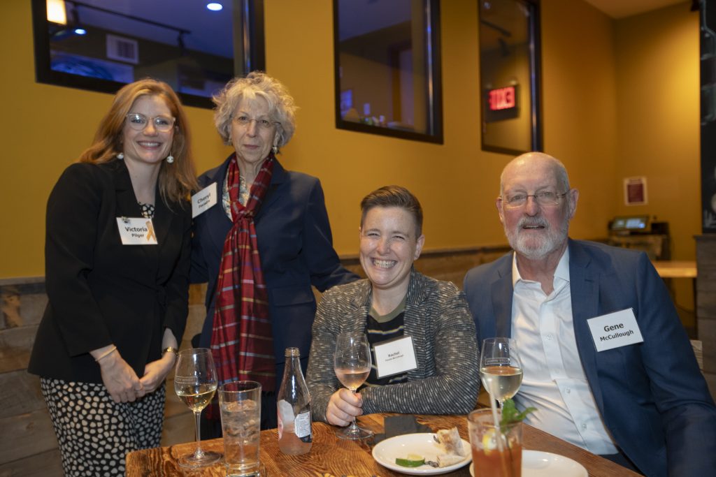 DRA's Victoria Pilger posing and smiling with three guests. Two of the guests are seated at a table. Three mirrors are hung on the wall behind them.