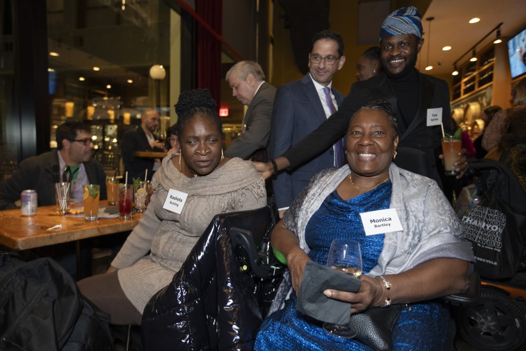 Four guests including DRA Advisory Board member Daniel Brown posing. Two guests are smiling. One guest is using a wheelchair, one guest is seated, and two guests are standing. One guest has his hand on another guest's shoulder. Other guests can be seen talking behind them.