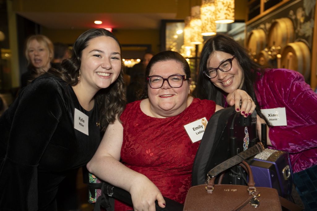 DRA Board member Emily Ladau posing and smiling with two guests. Emily is using a wheelchair. Another guest can be seen talking behind them. 