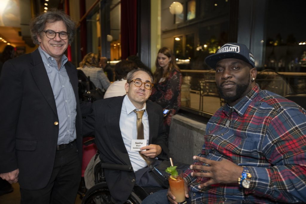 Three guests posing, two are using wheelchairs and the third is standing. Other guests are talking behind them and city lights can be seen through the windows.