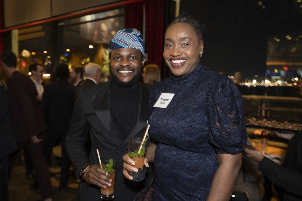 Two guests posing and smiling while other guests talk behind them. City lights can be seen through the window.