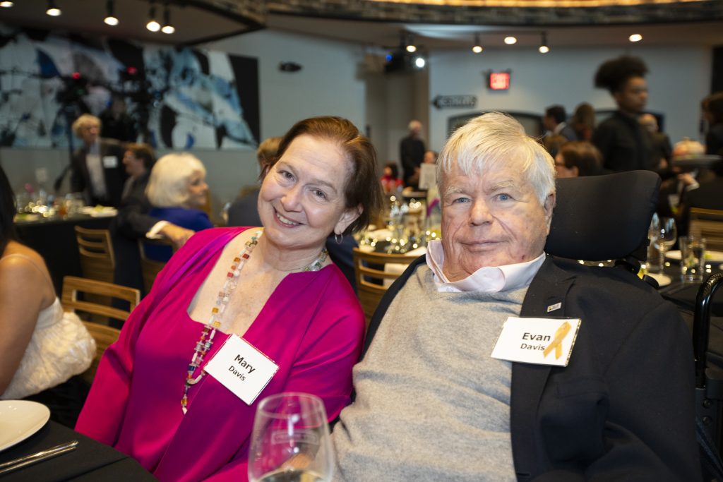 DRA Board member Evan Davis posing and smiling with another seated guest while using his wheelchair. Other guests are seated in the background.