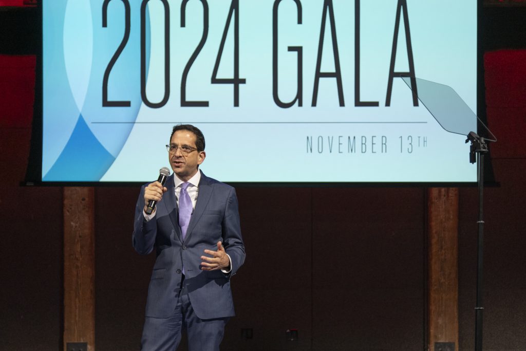 DRA Advisory Board member Daniel Brown on stage speaking into a microphone he is holding. The screen behind him says "2024 Gala, November 13th."