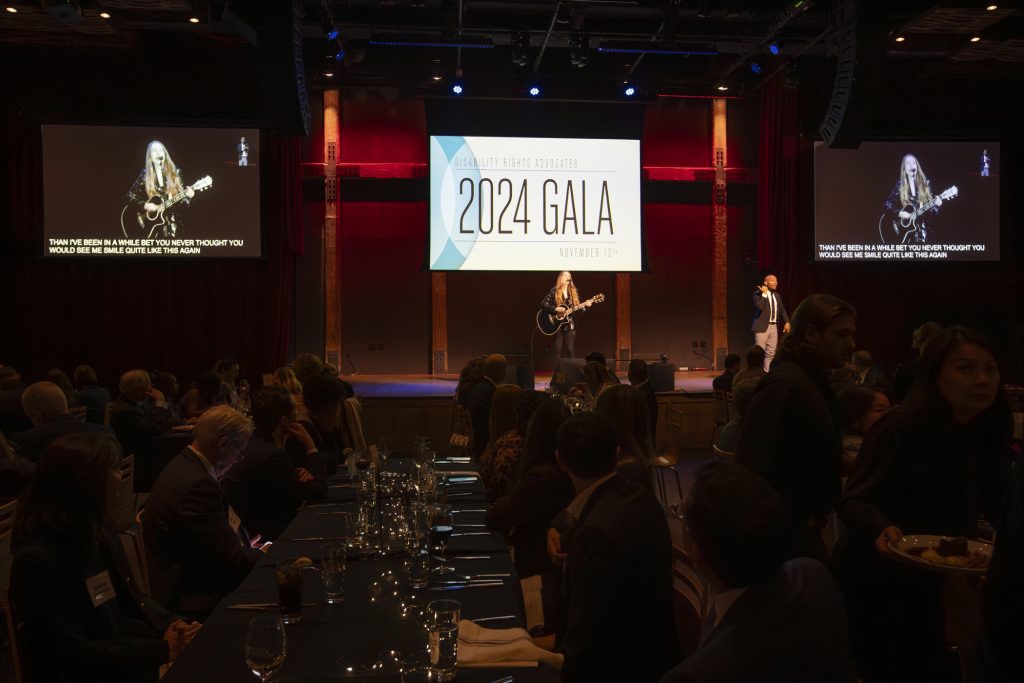 Special guest Stress Dolls playing guitar and singing on stage while seated guests watch. An ASL interpreter is on stage with her. Two screens on the sides of the stage show the performance and the main screen says "2024 Gala."
