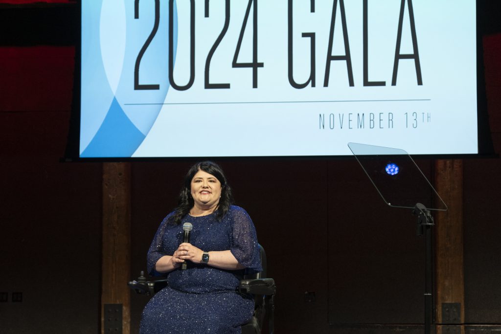 DRA's Rebecca Williford on stage speaking into a microphone she is holding while using her wheelchair. The screen behind her says "2024 Gala, November 13th."