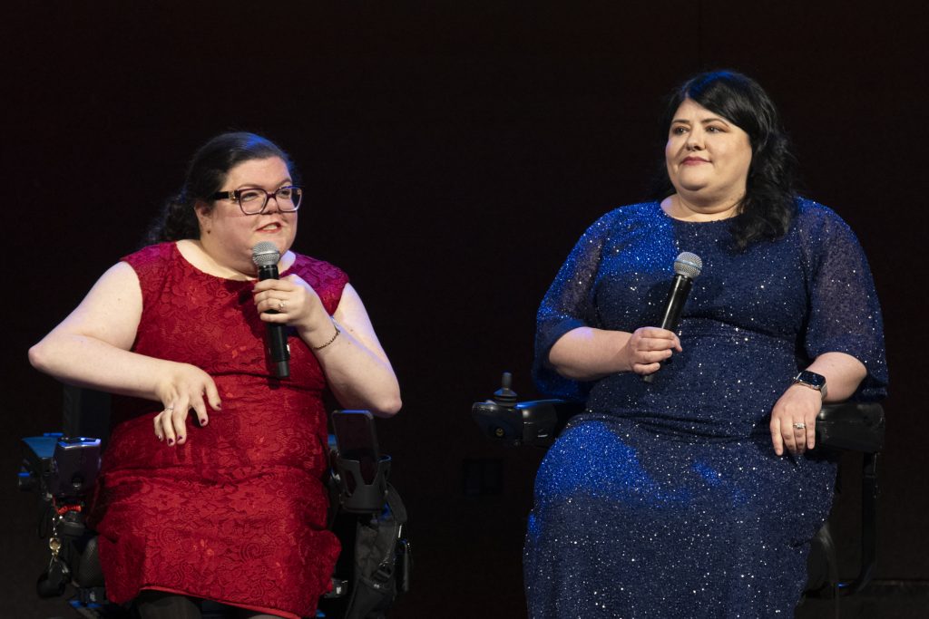 DRA's Rebecca Williford and DRA Board member Emily Ladau on stage holding microphones. Emily is speaking into her microphone, both are using wheelchairs.