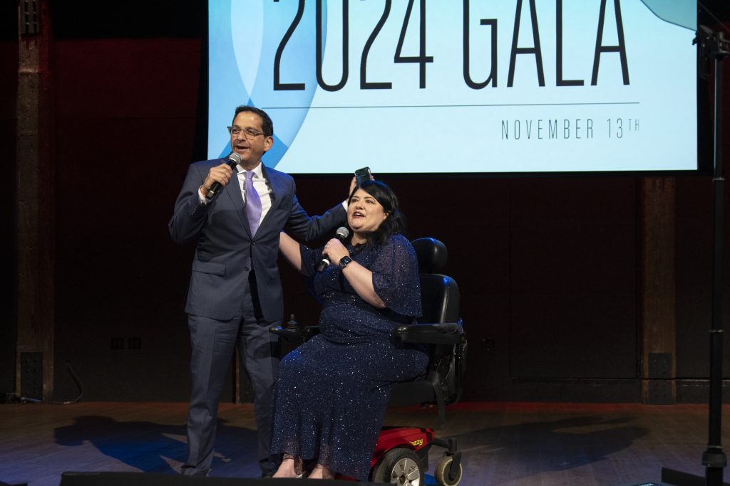 DRA Advisory Board member Daniel Brown and DRA's Rebecca Williford both speaking into the microphones they are holding on stage. Daniel has a phone in his hand and Rebecca has her arm around Daniel while using her wheelchair. The screen behind them says "2024 Gala, November 13th."