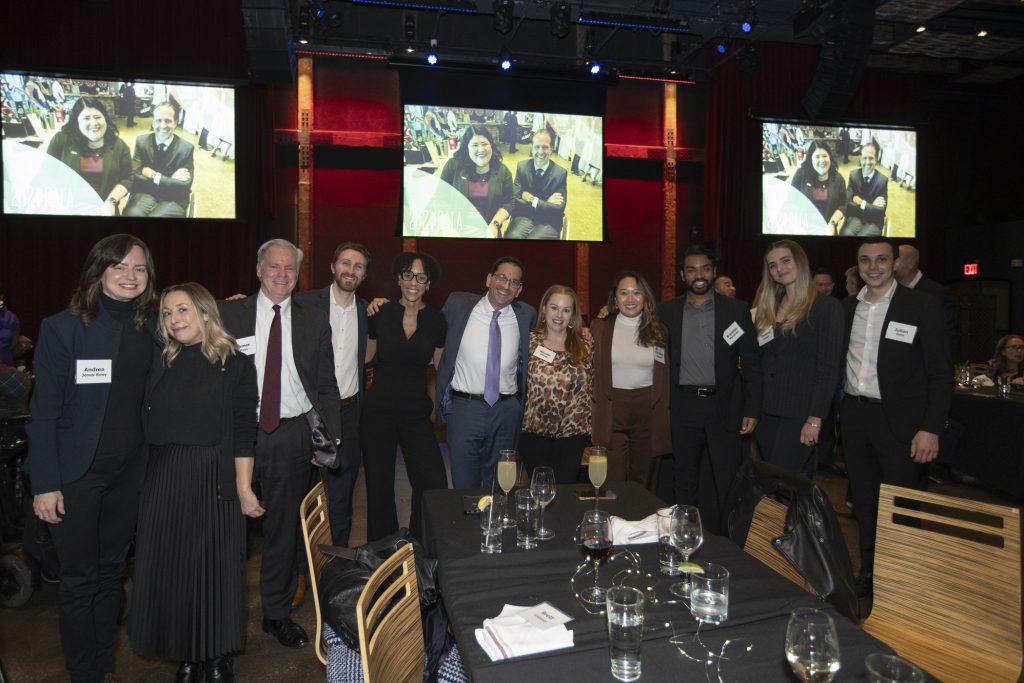 DRA Advisory Board member Daniel Brown of Sheppard Mullin posing with ten other Sheppard Mullin guests, all are smiling and have arms around each other. Three screens behind them on the wall show a picture of DRA's Rebecca Williford and honoree Yannick Benjamin.