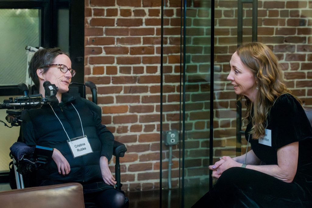 DRA Board member Cristina Rubke speaking with DRA's Kate Hamilton in front of a brick wall. Cristina is using her wheelchair and Kate is sitting. 