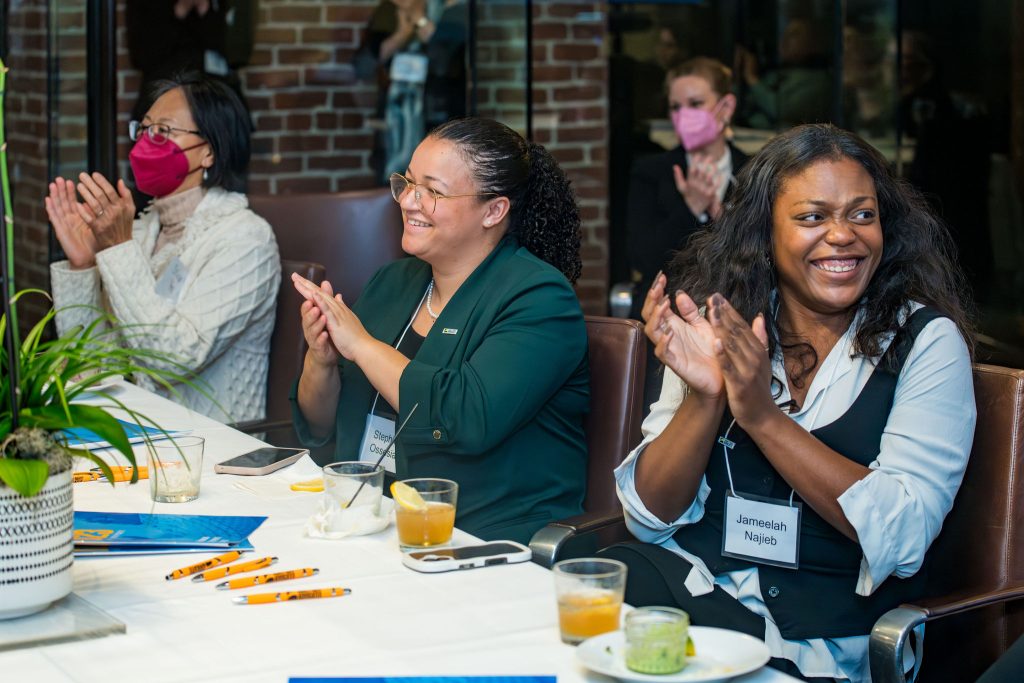 DRA's Stephie Ossesia, DRA's Jameelah Najieb, and a guest clapping and smiling from their seats as they watch the on-screen program. Other guests are clapping behind them.