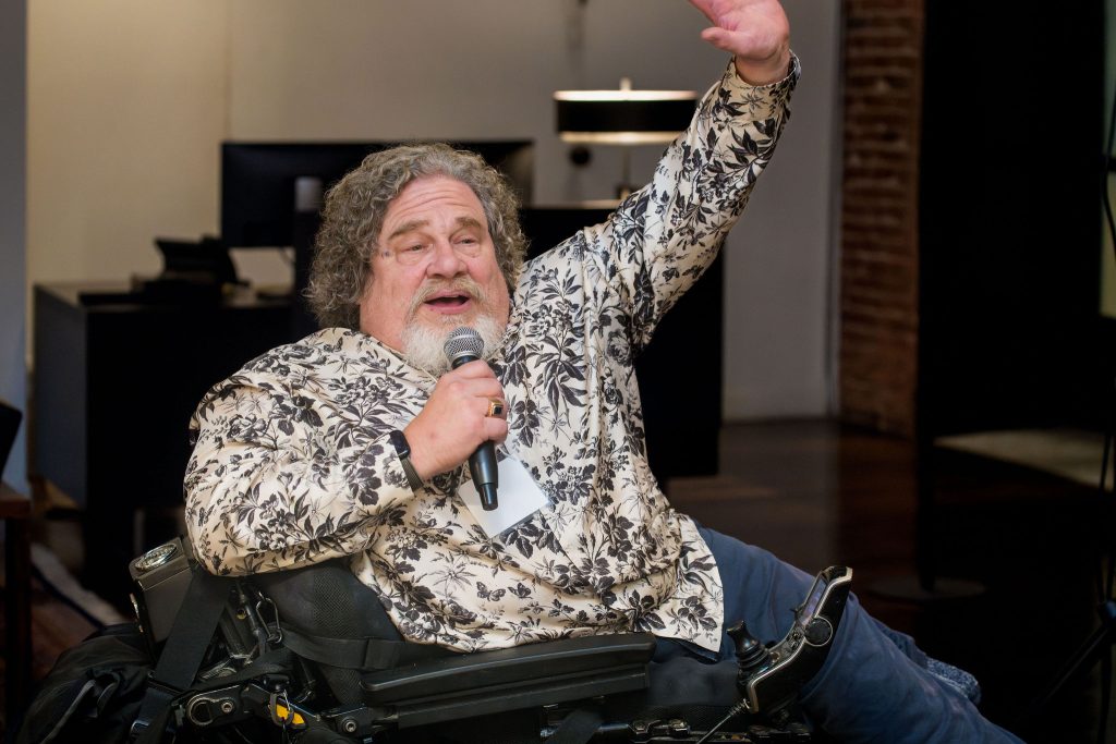 Special guest Jim LeBrecht raises his hand while speaking into a microphone from his wheelchair.