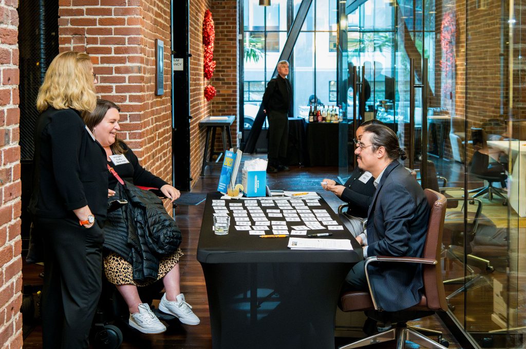 DRA's Shawna Parks, DRA Board member Morgan Duffy-Granville, DRA's Chris Maccarone, and DRA's Christin Mills speaking with each other. Chris and Christin are seated at a table with nametags spread on it. Shawna is standing and Morgan is using her wheelchair. Brick walls and large windows are in the background.