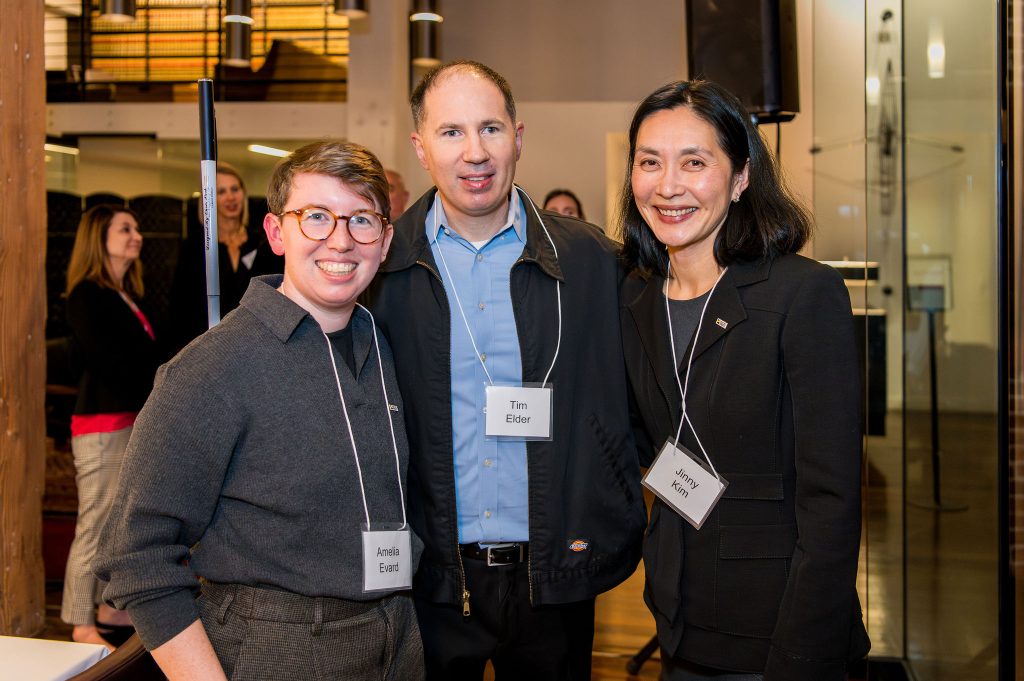 DRA's Amelia Evard and DRA's Jinny Kim posing and smiling with a guest holding his white cane. Other guests are in the background.