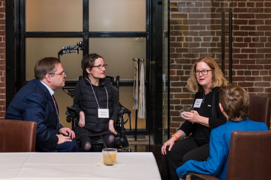DRA's Scott Gordon, DRA Board member Cristina Rubke, DRA's Shawna Parks, and a guest sitting and talking in front of a brick wall. Cristina is using her wheelchair.