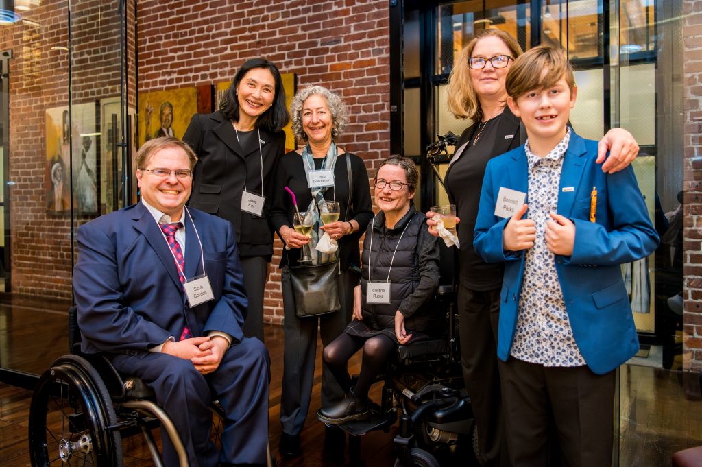 DRA's Scott Gordon, DRA's Jinny Kim, DRA Advisory Board member Linda Dardarian, DRA Board member Cristina Rubke, DRA's Shawna Parks, and a guest posing and smiling in front of a brick wall. Scott and Cristina are using their wheelchairs.