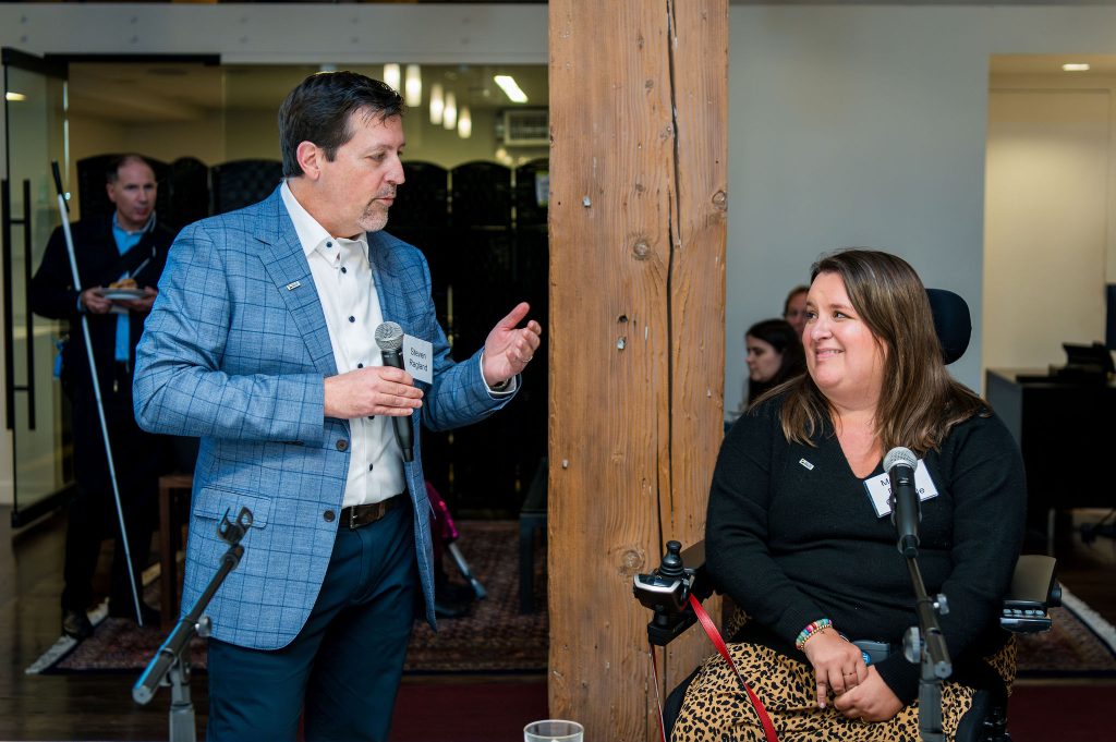 DRA Board member Steven Ragland gesturing with his hand towards DRA Board member Morgan Duffy-Granville next to him while speaking into the microphone he is holding. Other guests can be seen behind them.