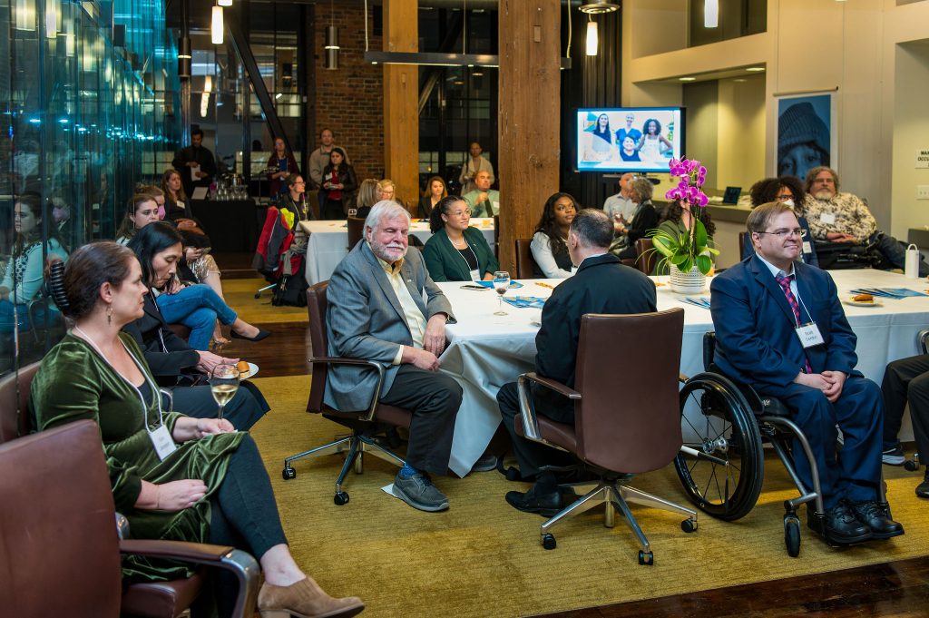 Many guests, mostly seated with some people standing or using wheelchairs, watching the on-screen program currently displaying DRA's Rebecca Williford.