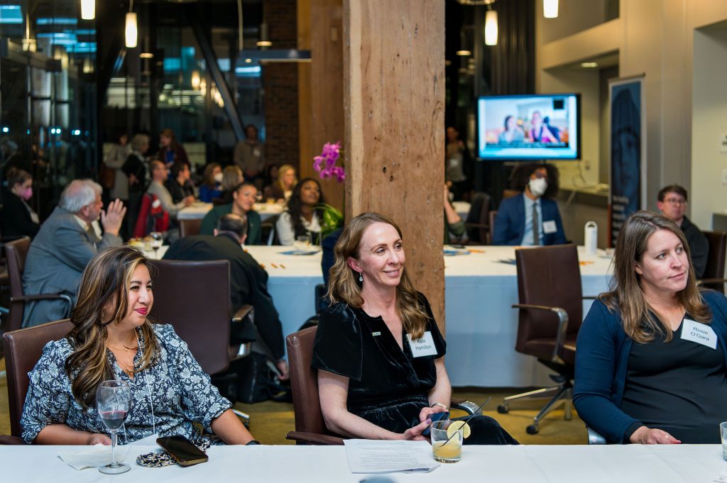 DRA Advisory Board member Natalie Aliga, DRA's Kate Hamilton, and another guest watching the on-screen program from their seats at a table, currently displaying DRA Board member Shane Burcaw and his wife Hannah Burcaw. Many other guests are behind them also watching the program.
