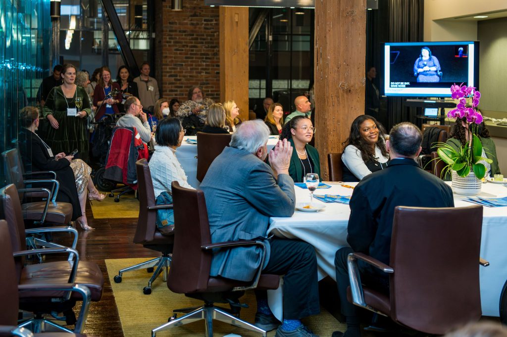Many guests, mostly seated with some people standing or using wheelchairs, watching the on-screen program currently displaying DRA's Rebecca Williford.