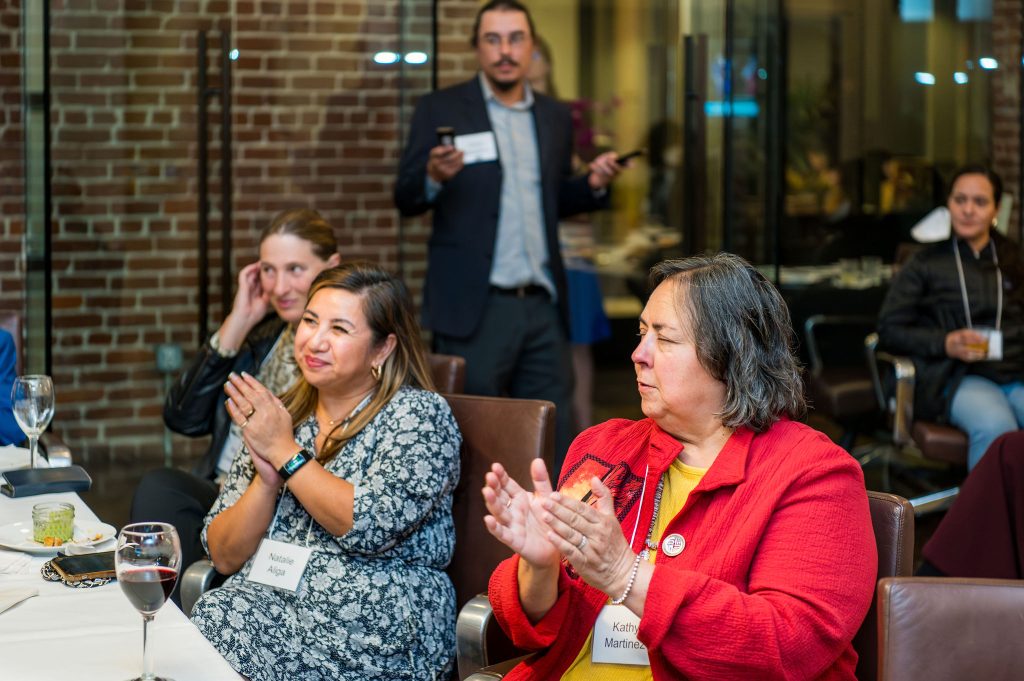 DRA Advisory Board member Anya Emerson, DRA Advisory Board member Natalie Aliga, and DRA Board member Kathy Martinez clapping from their seats as they watch the on-screen program. DRA's Chris Maccarone is standing in the background holding two remote controls and another guest is seated nearby.