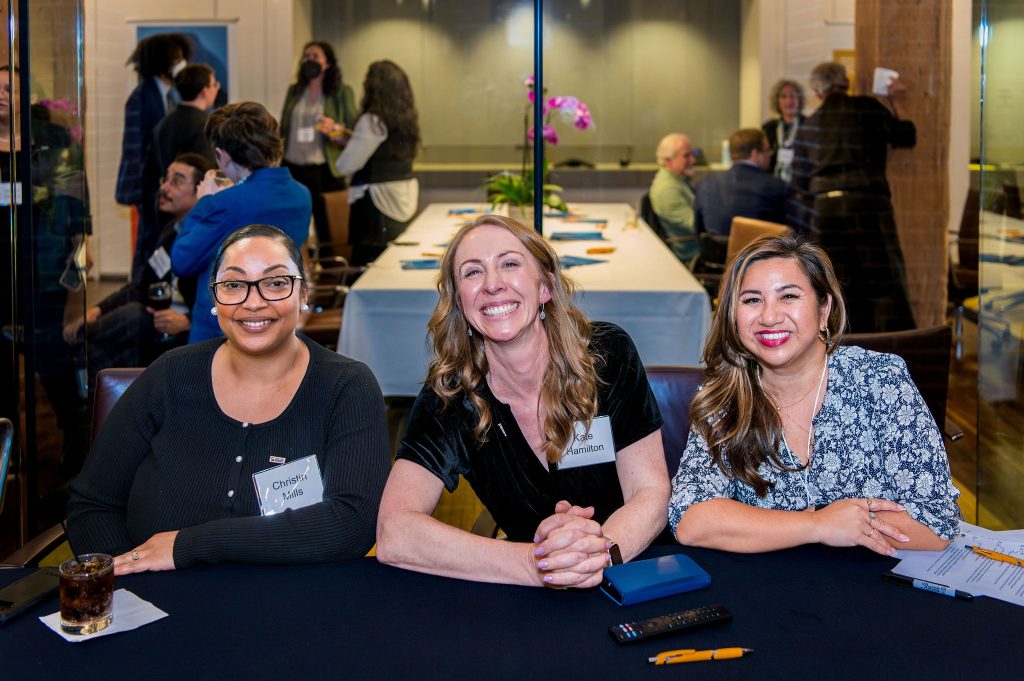 DRA's Christin Mills, DRA's Kate Hamilton, and DRA Advisory Board member Natalie Aliga posing and smiling while sitting at a table. Other guests can be seen talking amongst themselves in groups behind them.